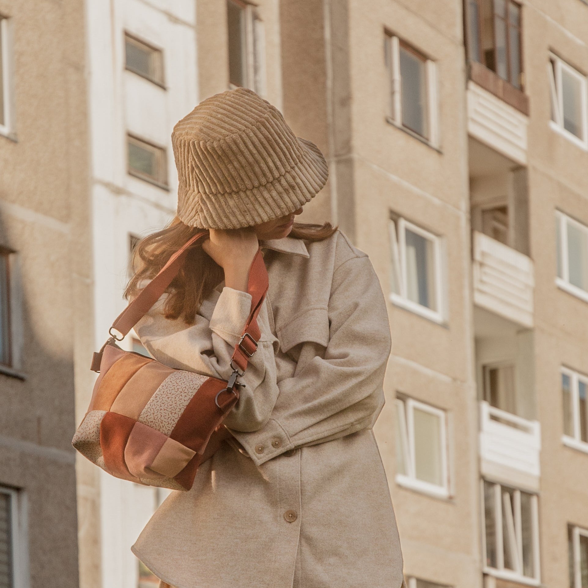 A person wearing a beige coat and a matching bucket hat, carrying a beige, red, and brown shoulder bag.