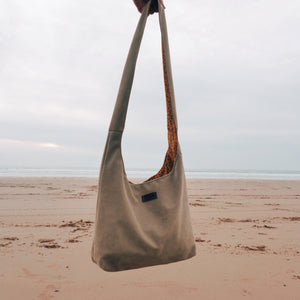Beige shoulder bag held by a hand on a sandy beach with a cloudy sky.