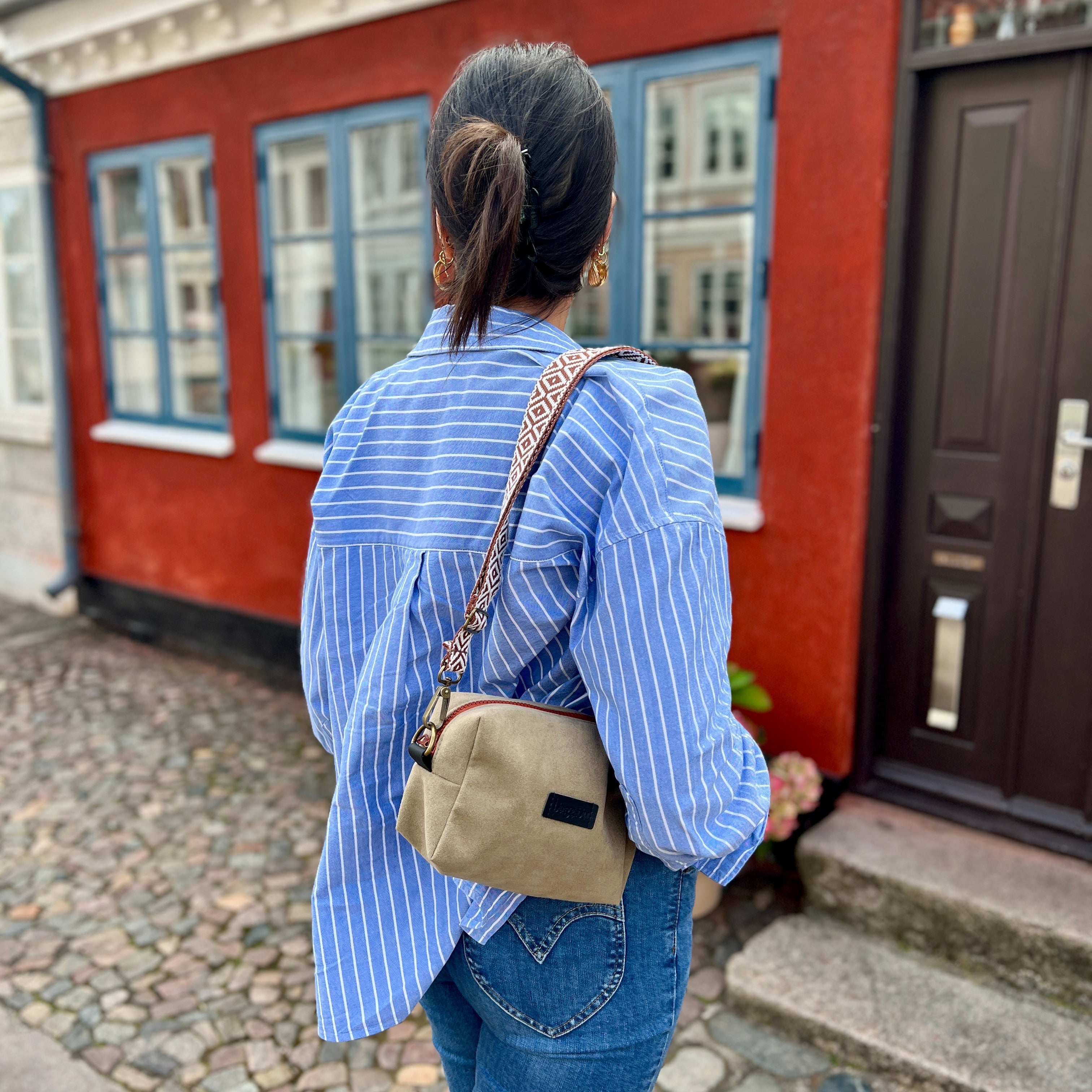 Person wearing a blue striped shirt and jeans with a beige crossbody bag, standing in front of a red building.