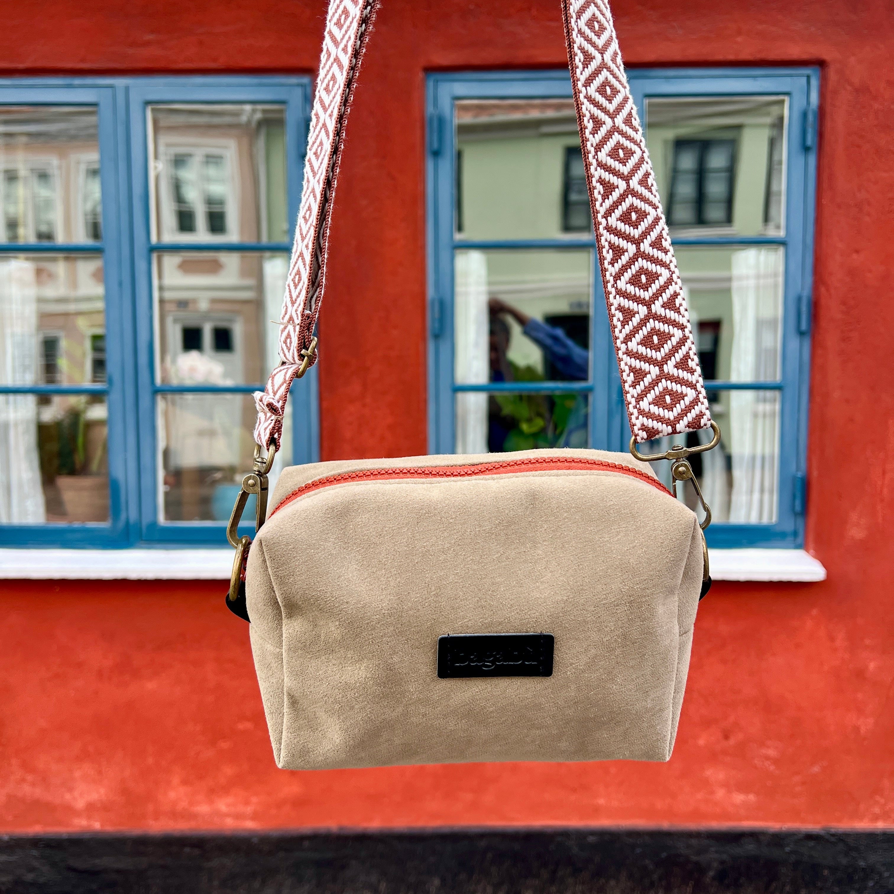 Beige shoulder bag with a patterned strap against a red building with blue windows.
