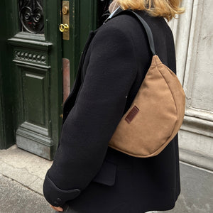 A person wearing a black coat and blue jeans is carrying a sand-colored banana bag over their shoulder.