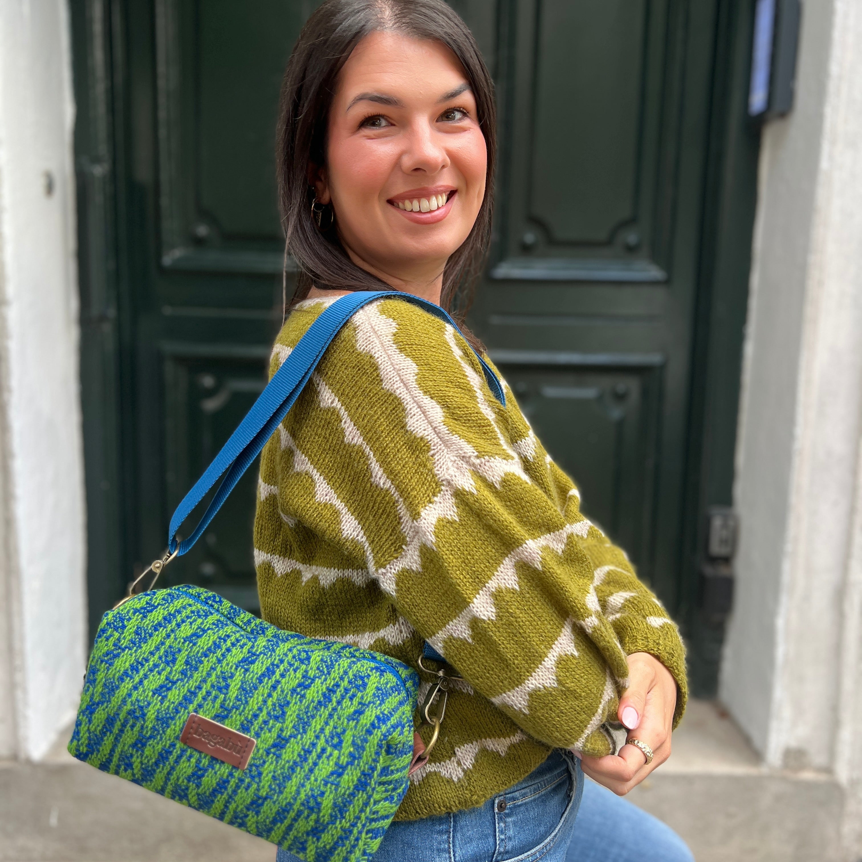 Woman wearing a green patterned sweater with a blue and green bag, standing in front of a dark door.