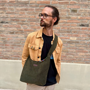 Man wearing a tan jacket and holding a green bag against a brick wall.