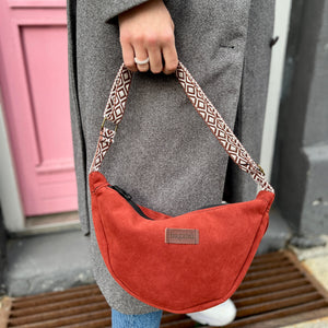 A person holding a cherry red banana-shaped handbag with a patterned strap.