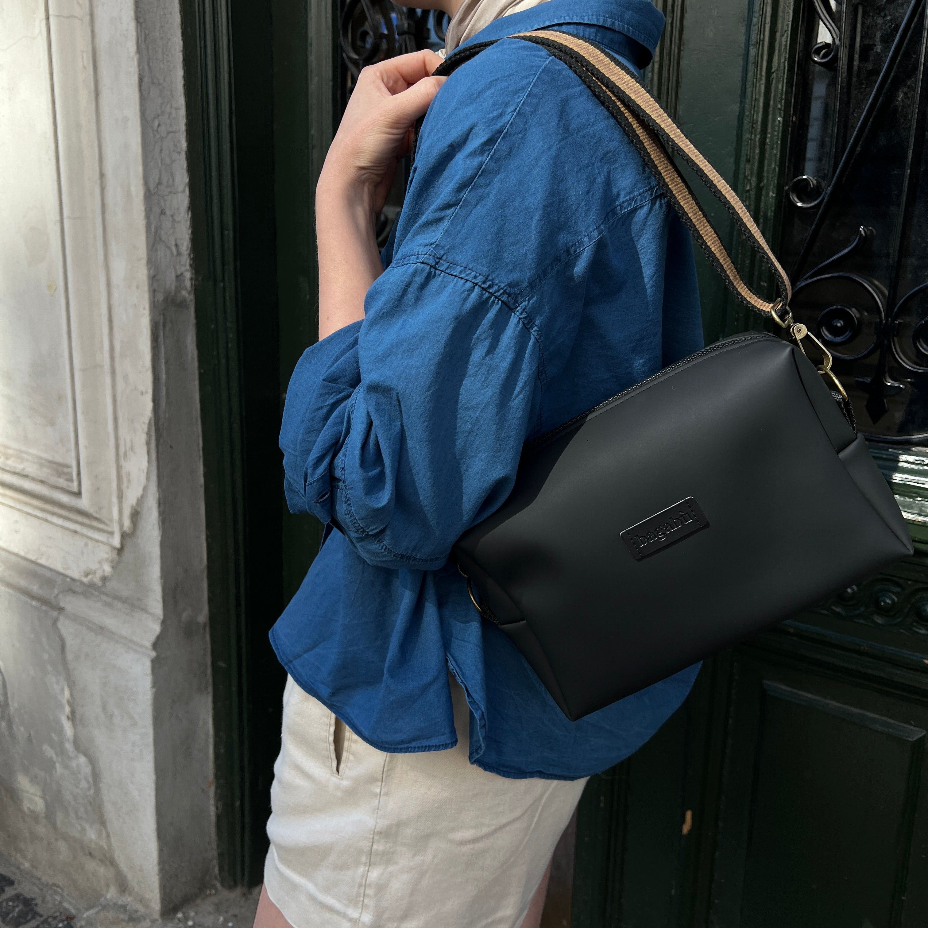 A person wearing a blue shirt and white skirt is carrying a black shoulder bag with a tan shoulder strap over their shoulder.