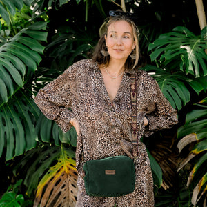 A person wearing a leopard print outfit is standing in front of green foliage, carrying a green Moss Alcantara bag.