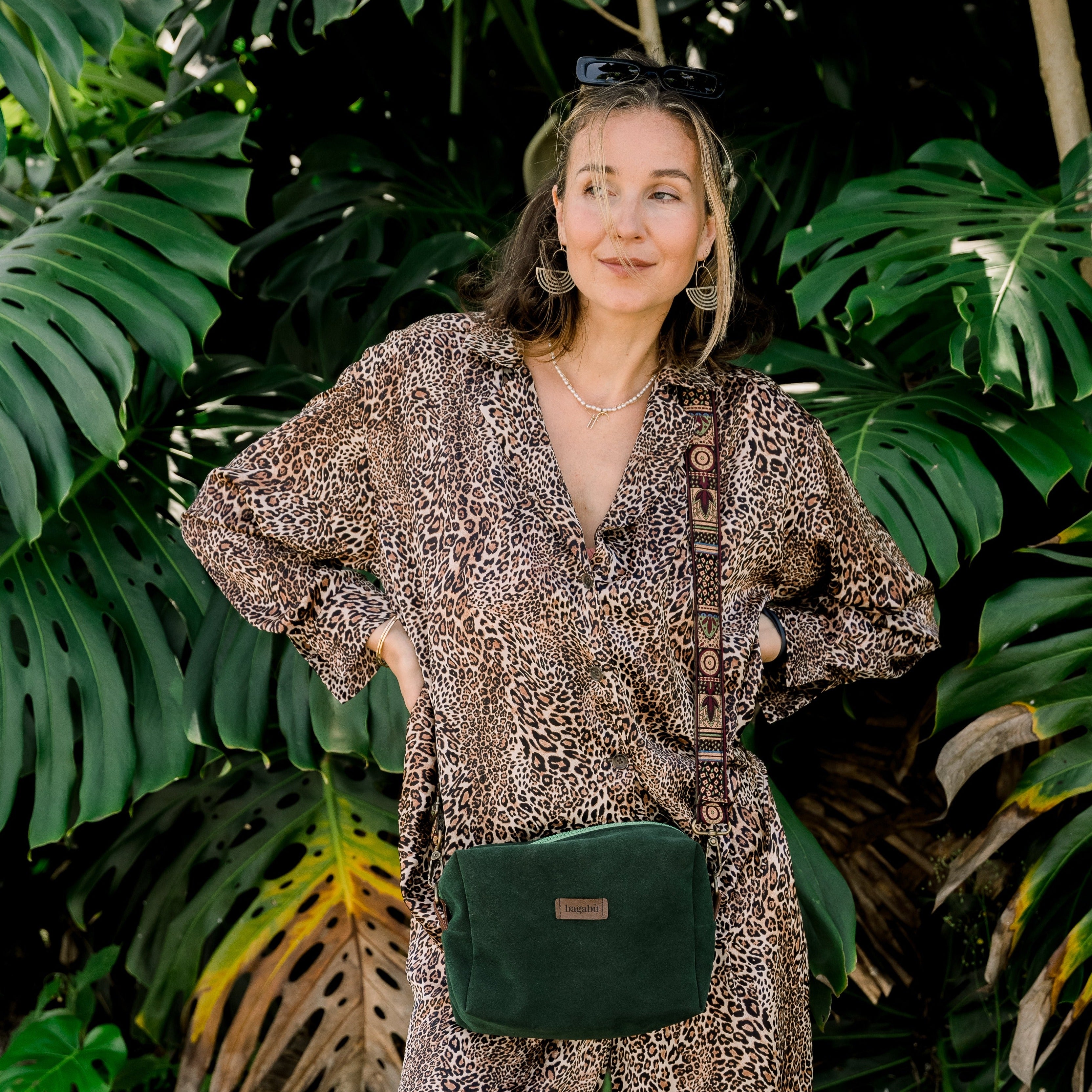 A person wearing a leopard print outfit is standing in front of green foliage, carrying a green Moss Alcantara bag.