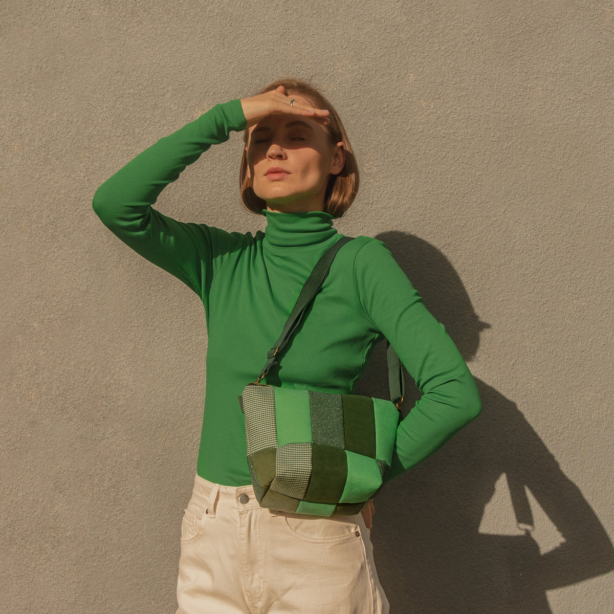 A person standing with a green turtleneck and green bag, with upcycled green and blue textures on the bag.