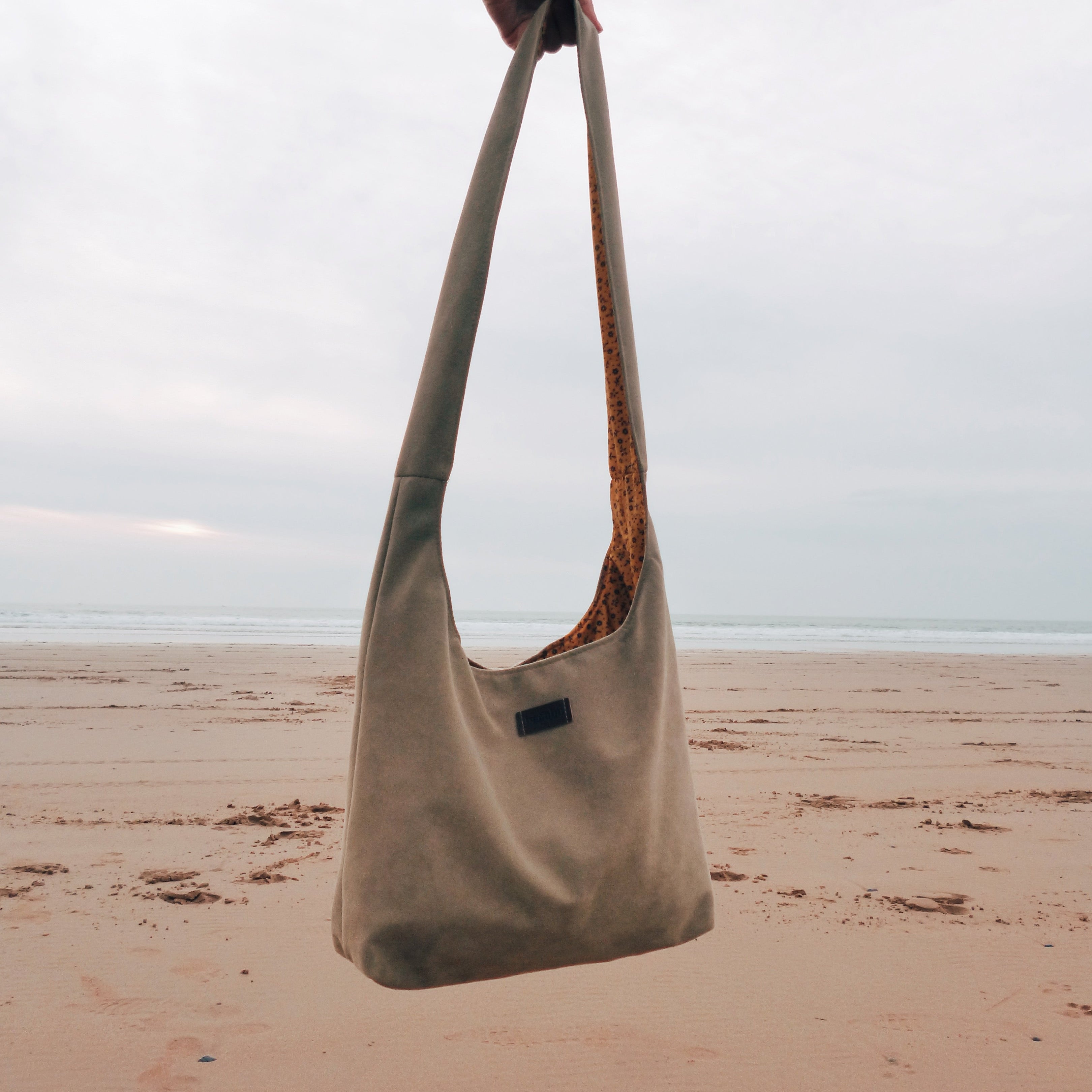 Beige shoulder bag held by a hand on a sandy beach with a cloudy sky.