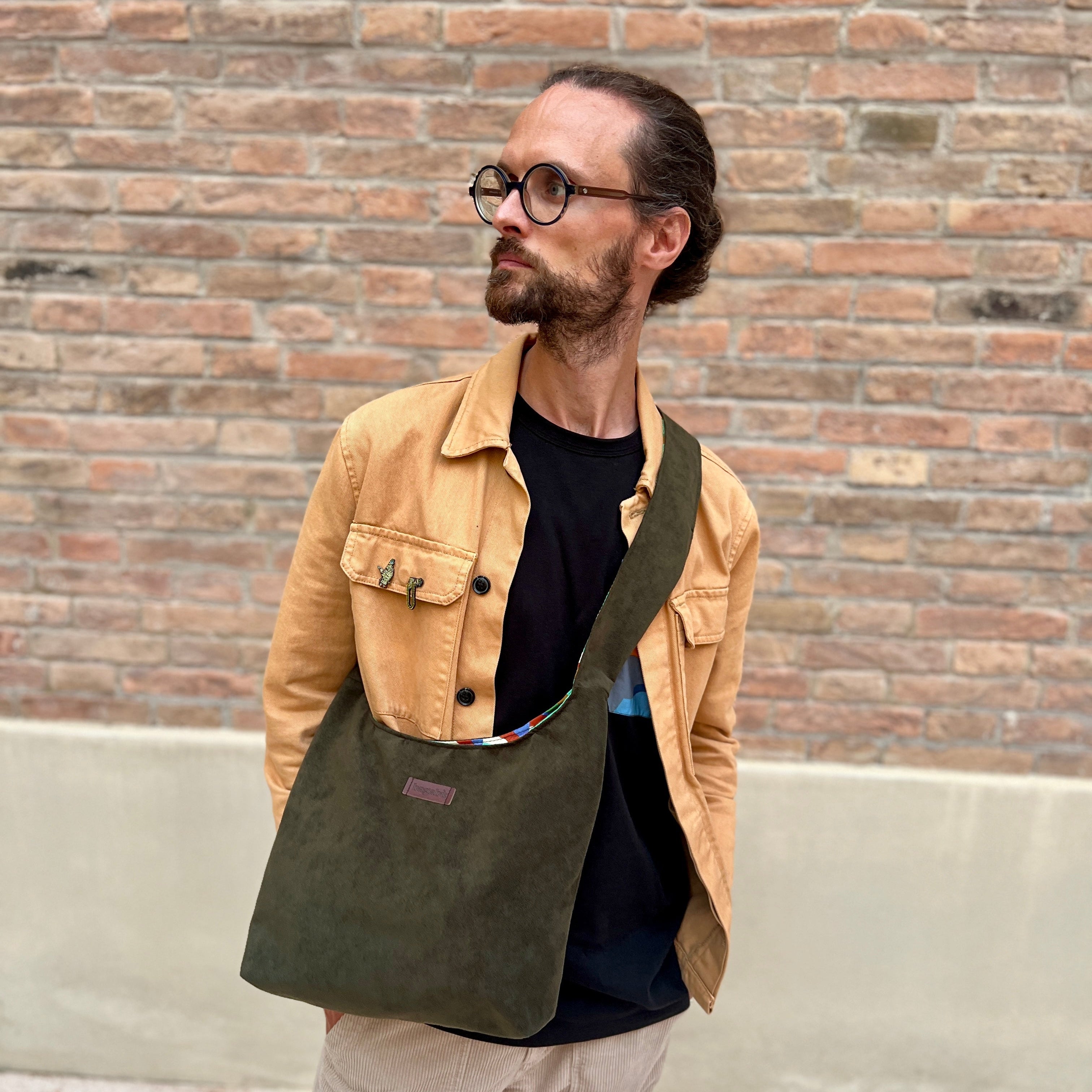 Man wearing a tan jacket and holding a green bag against a brick wall.