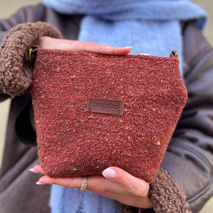 Person holding a brown textured pouch with a brand label, wearing a blue scarf and brown coat.